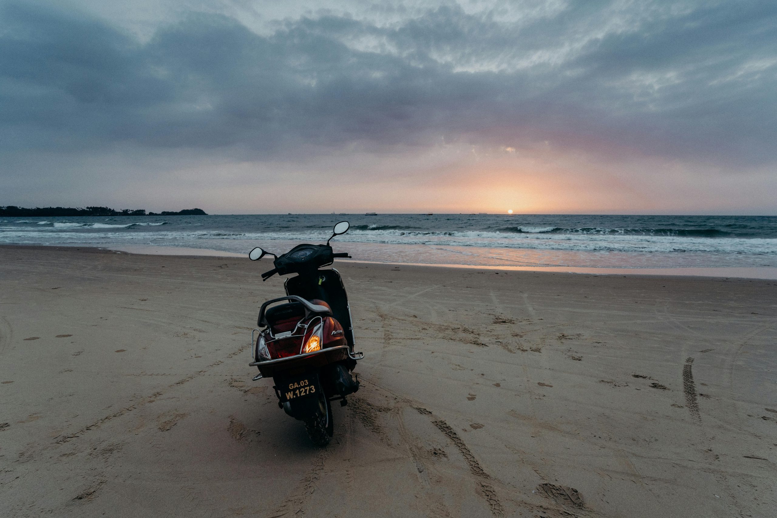 A motorcycle is parked on a tranquil Goa beach as the sun sets over the ocean. Perfect for travel inspiration.